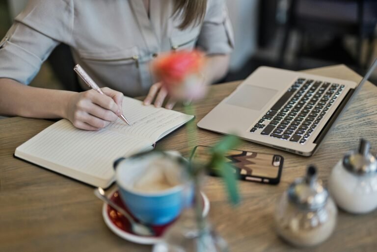 Blog Woman writing in a notebook with a laptop and coffee cup on a desk. Ideal for workspace inspiration.