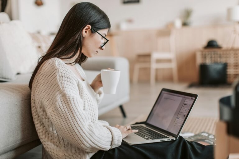 Blog Side view of female freelancer in warm sweater and eyeglasses drinking tea from white ceramic cup while sitting on floor near sofa with netbook on legs while creating document for remote work project