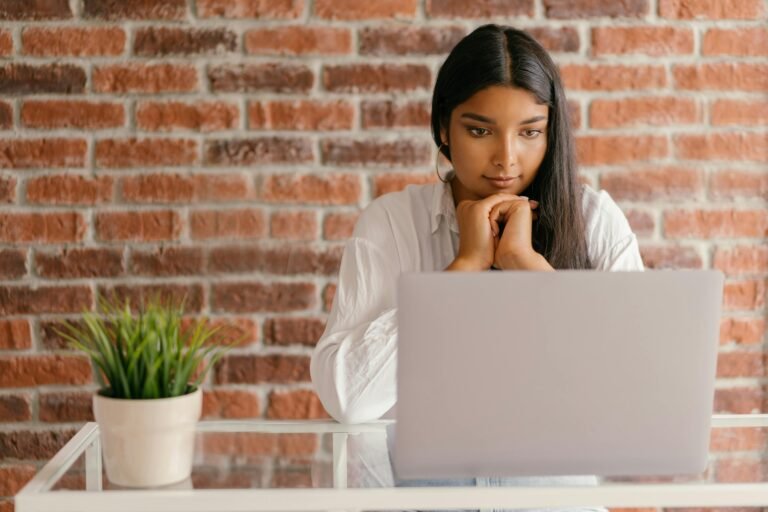 Blog Young woman deeply focused on her laptop studying indoors against a brick wall backdrop.